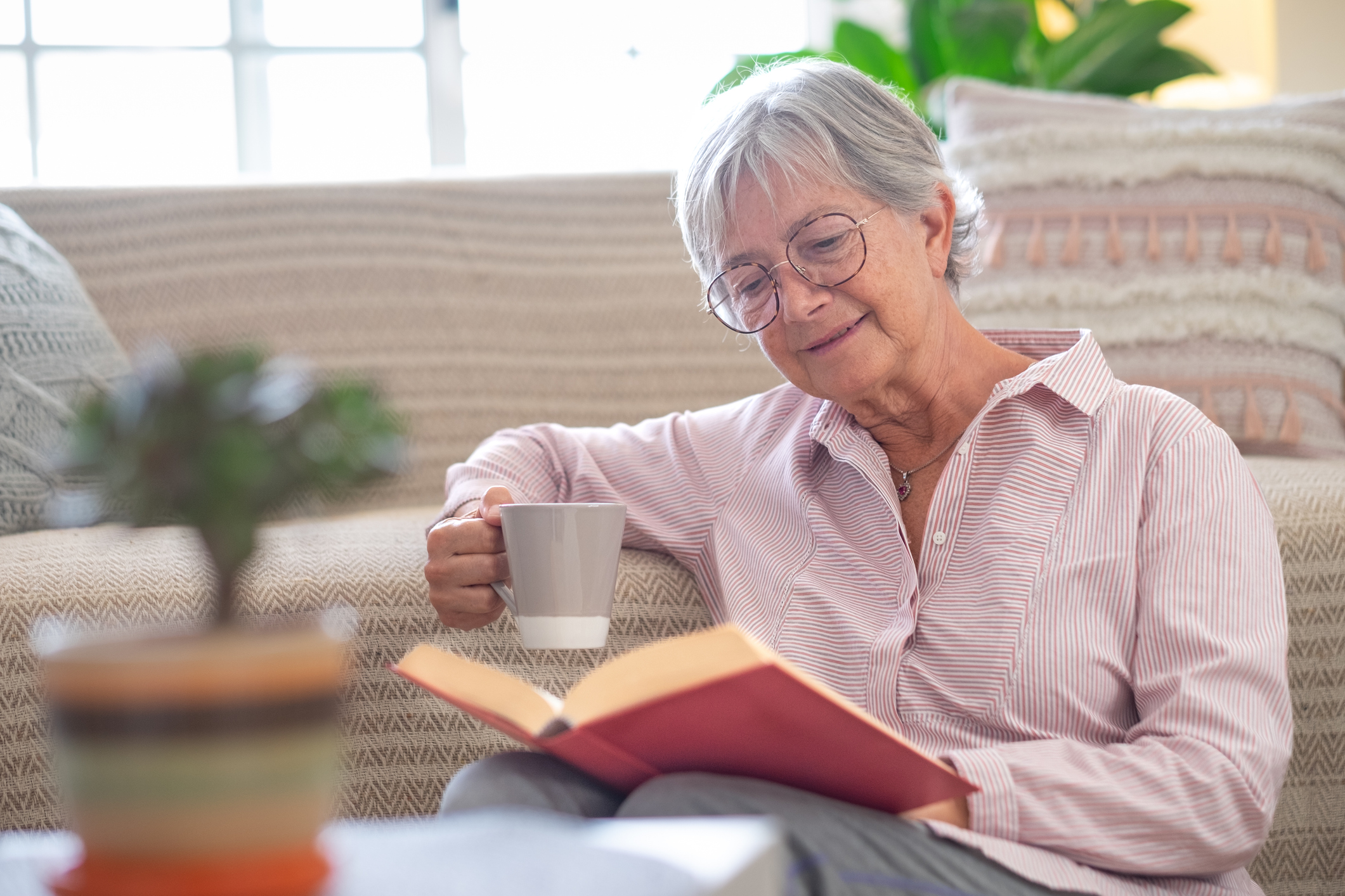 Smiling attractive senior woman relaxing sitting on the floor reading a book holding a cup of coffee or tea. Caucasian mature lady studying, learning at home Smiling attractive senior woman relaxing sitting on the floor reading a book holding a cup of coffee or tea. Caucasian mature lady studying, learning at home
