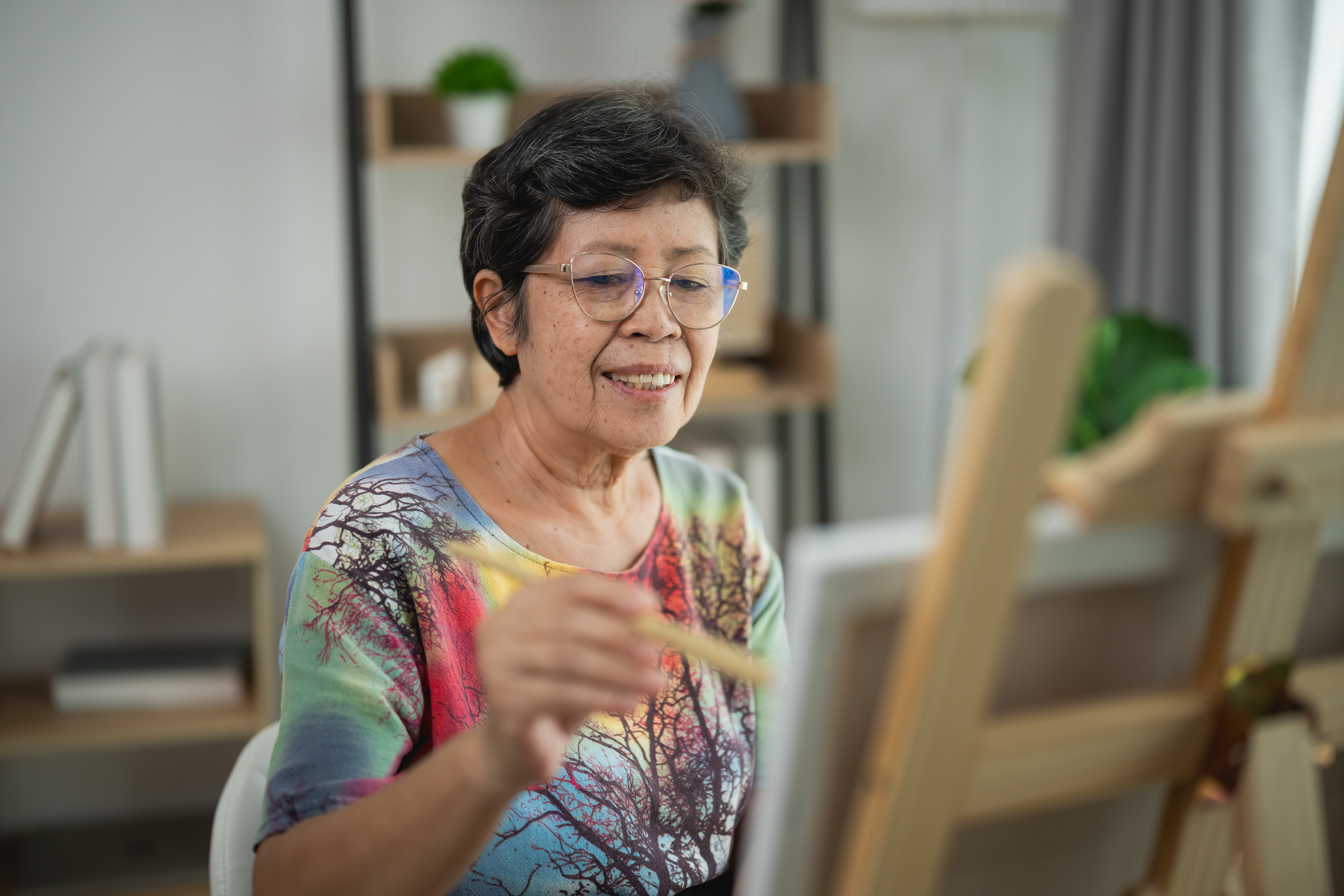 Senior woman painting on canvas indoors, enjoying art therapy, creating colorful masterpiece with brushes, smiling and focused in bright room