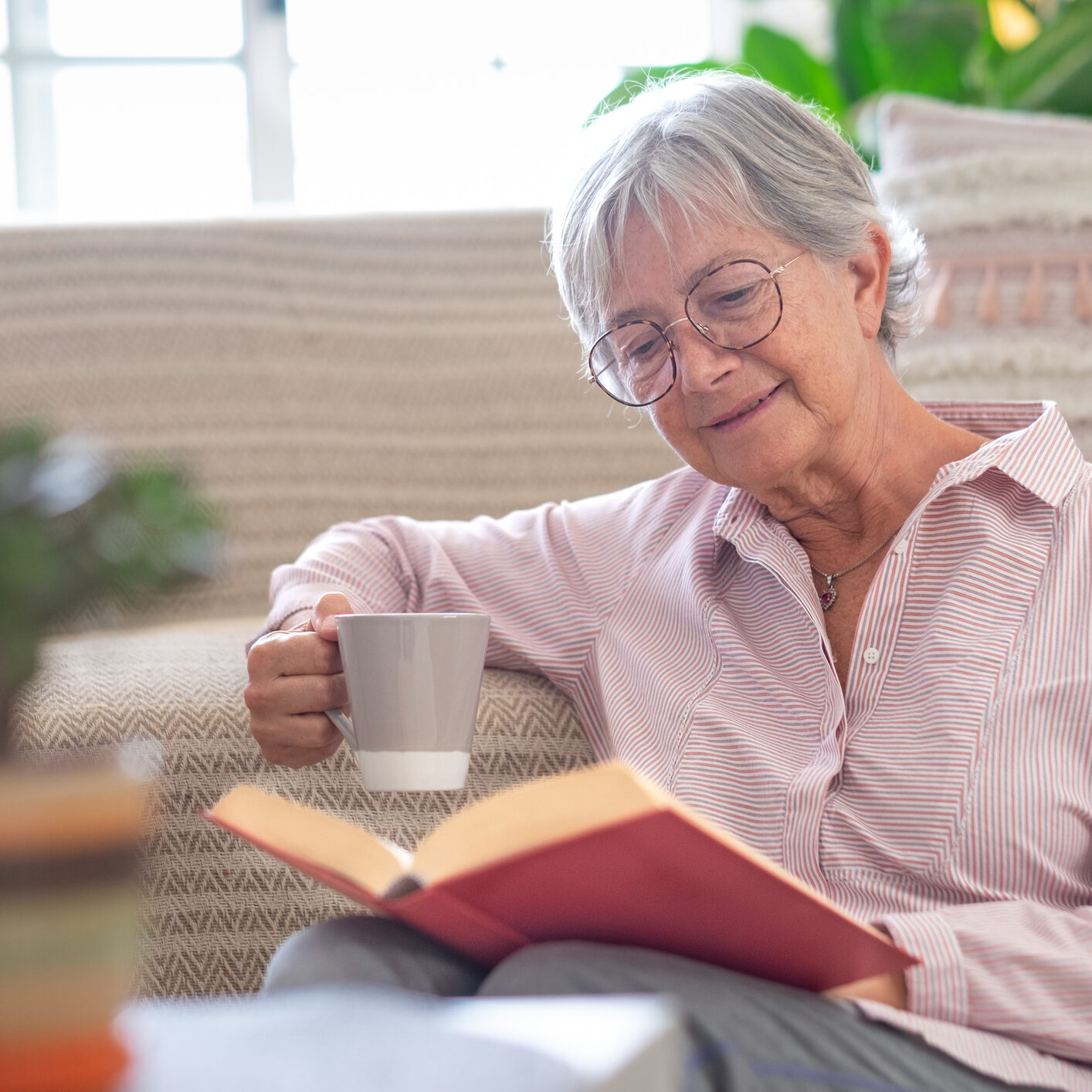 Smiling attractive senior woman relaxing sitting on the floor reading a book holding a cup of coffee or tea. Caucasian mature lady studying, learning at home